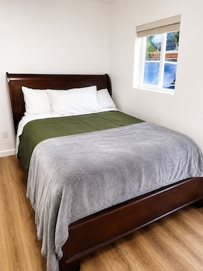 Una cama individual con cabecera de madera, colcha verde y gris, y ventana con vistas al cielo azul y árboles.