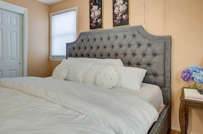 Un dormitorio con cabecera capitonada, ropa de cama blanca, una ventana con persianas y un florero en la pared con flores.