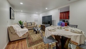 Un salón con sofá, una mesa de comedor con platos y un jarrón de flores, y una zona de cocina con armarios y televisor.