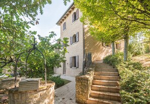 A two-story building with a brick facade, a stone staircase, and a garden area with trees and shrubs. - Villa Belvedere San Bartolo by PosarelliVillas (Montefiore Conca)