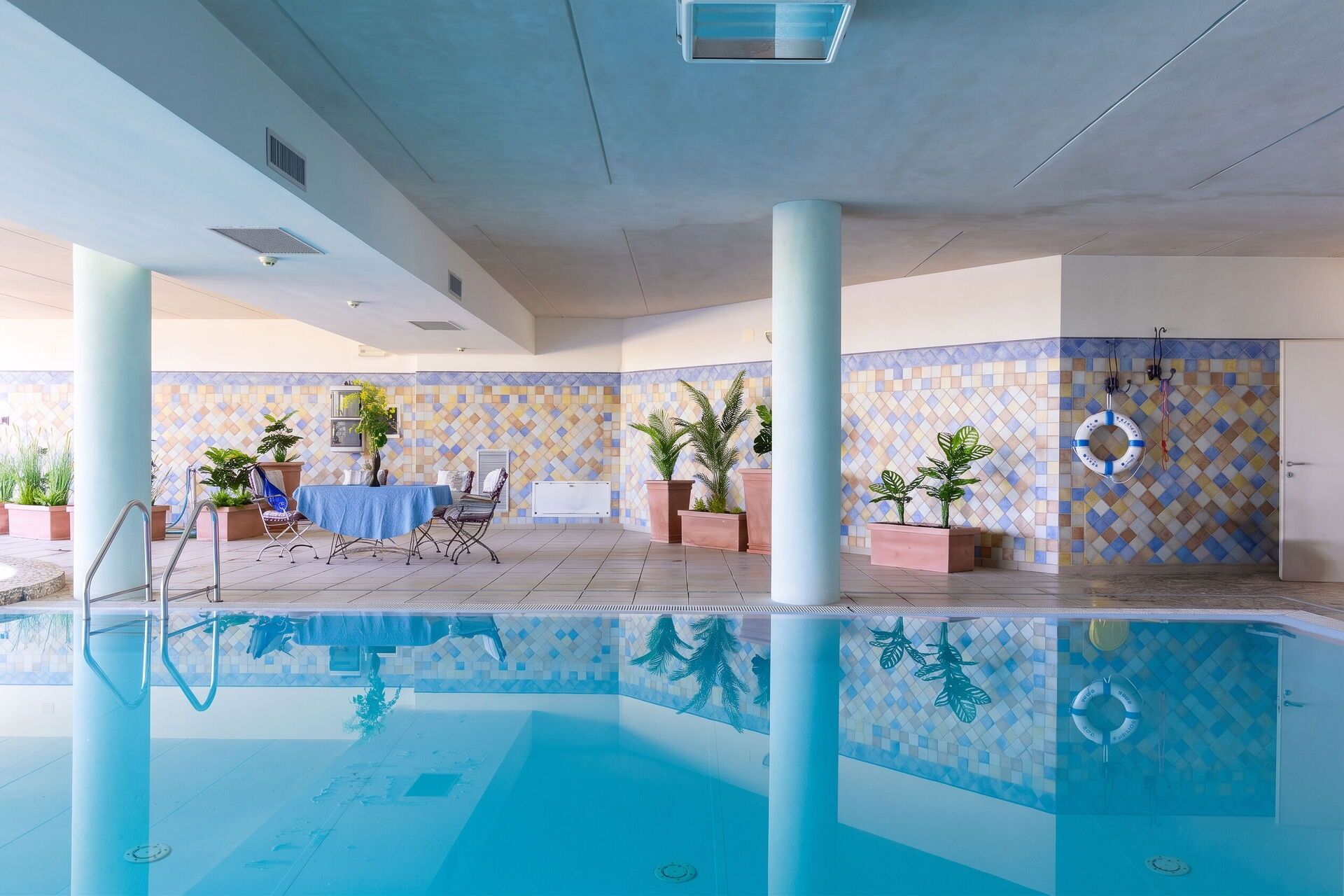 A swimming pool area with a tiled wall, a table with chairs, and potted plants.