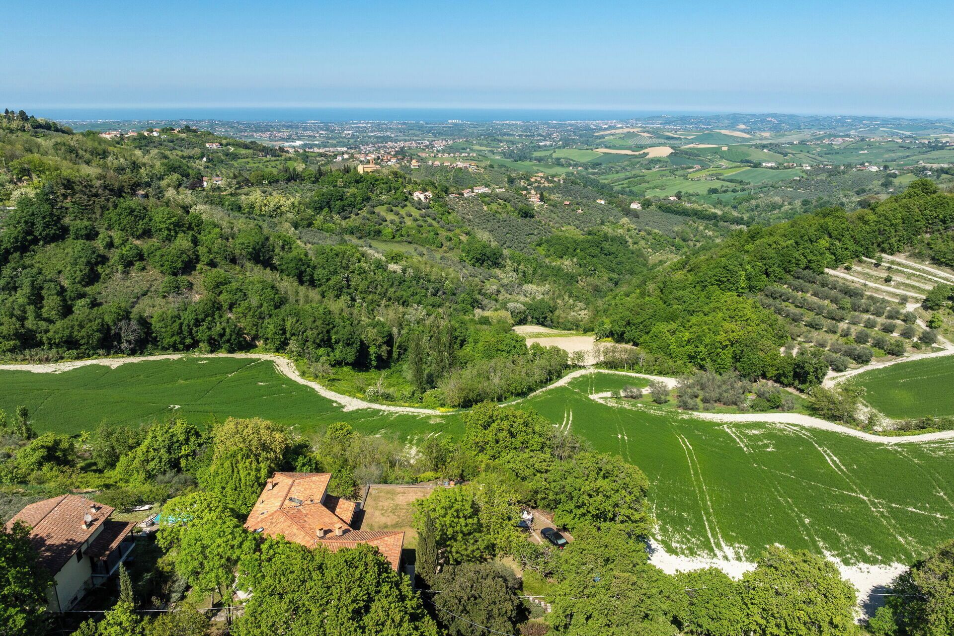 A rural landscape with terraced fields, a small building with a tiled roof, and a distant view of a town by the sea.