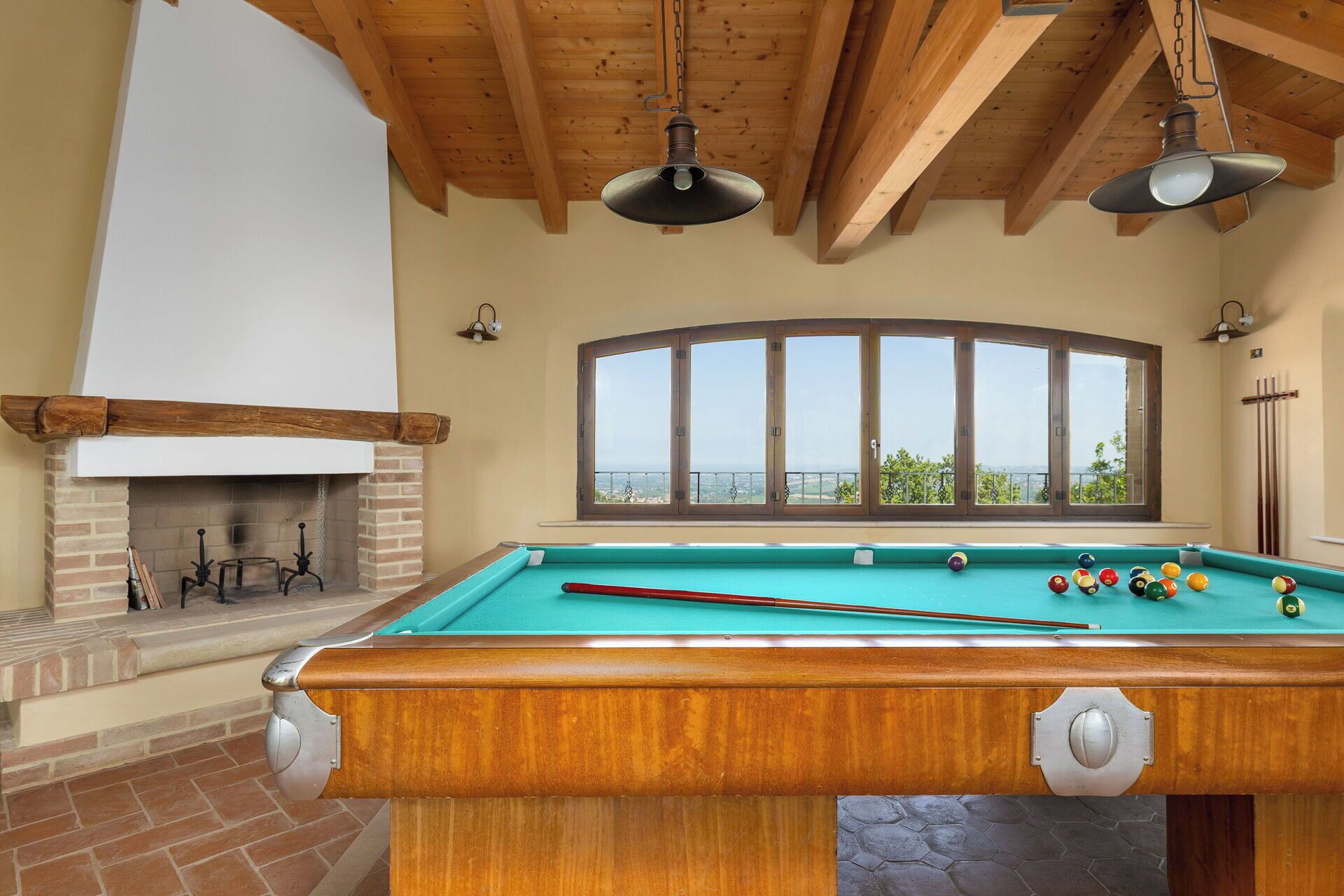 A pool table with a view of the outdoors, a fireplace, and wooden beams on the ceiling.