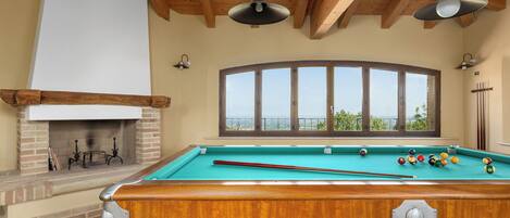 A pool table with a view of the outdoors, a fireplace, and wooden beams on the ceiling.