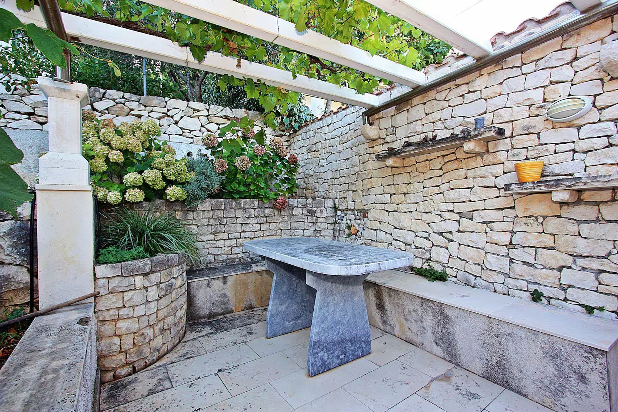 A stone wall with a built-in bench, a pergola with vines, and a stone shelf with a pot.