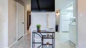 Um corredor leva a uma cozinha, com um respiradouro preto no teto, uma pequena mesa com uma planta e uma placa na parede.