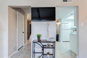 A hallway leading to a kitchen with a black ceiling vent, a small table with a plant, and a sign on the wall.