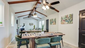 Uma sala de jantar com teto alto, vigas aparentes e iluminação pendente. Há cadeiras estofadas em verde ao redor de uma mesa de madeira.