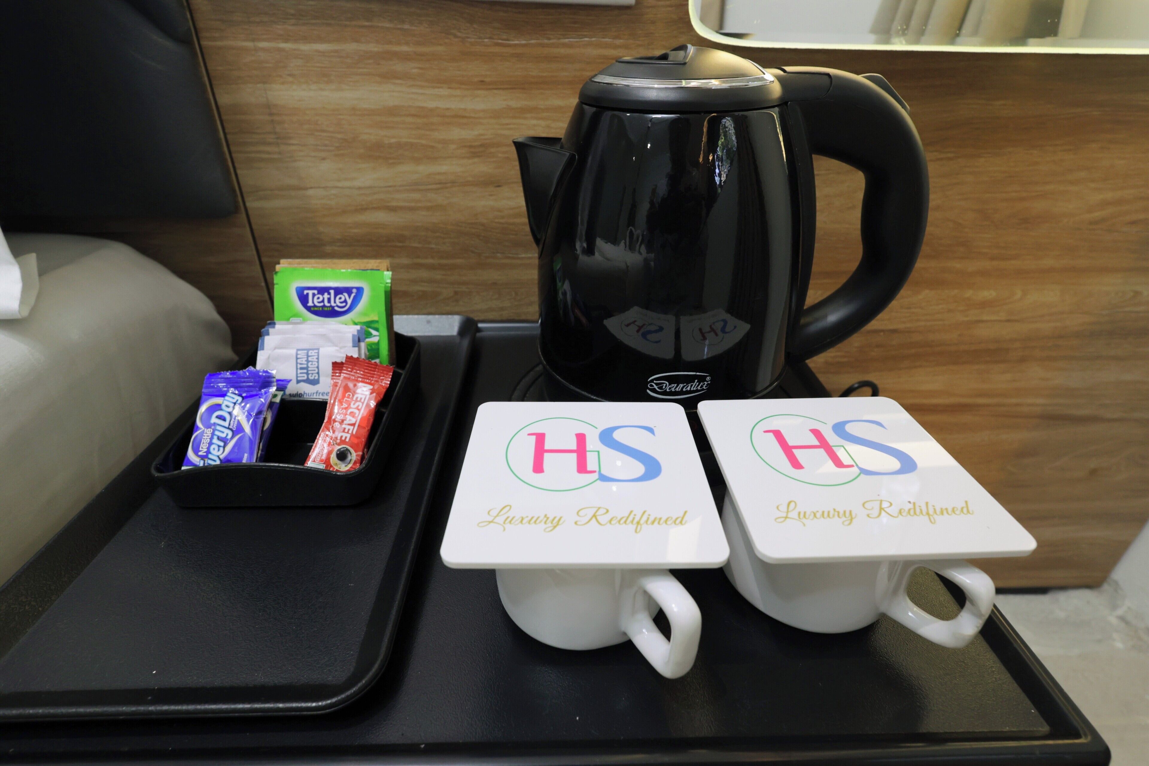 A tray with two white mugs labeled 'HS Luxury Redefined,' a black kettle, and some Tetley tea packets.