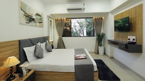 A hotel room with a bed, a TV, an air conditioner, a window with curtains, a plant, and a bedside table with a lamp and phone. - Chembur Homeotel - Gagal Home (Mumbai)