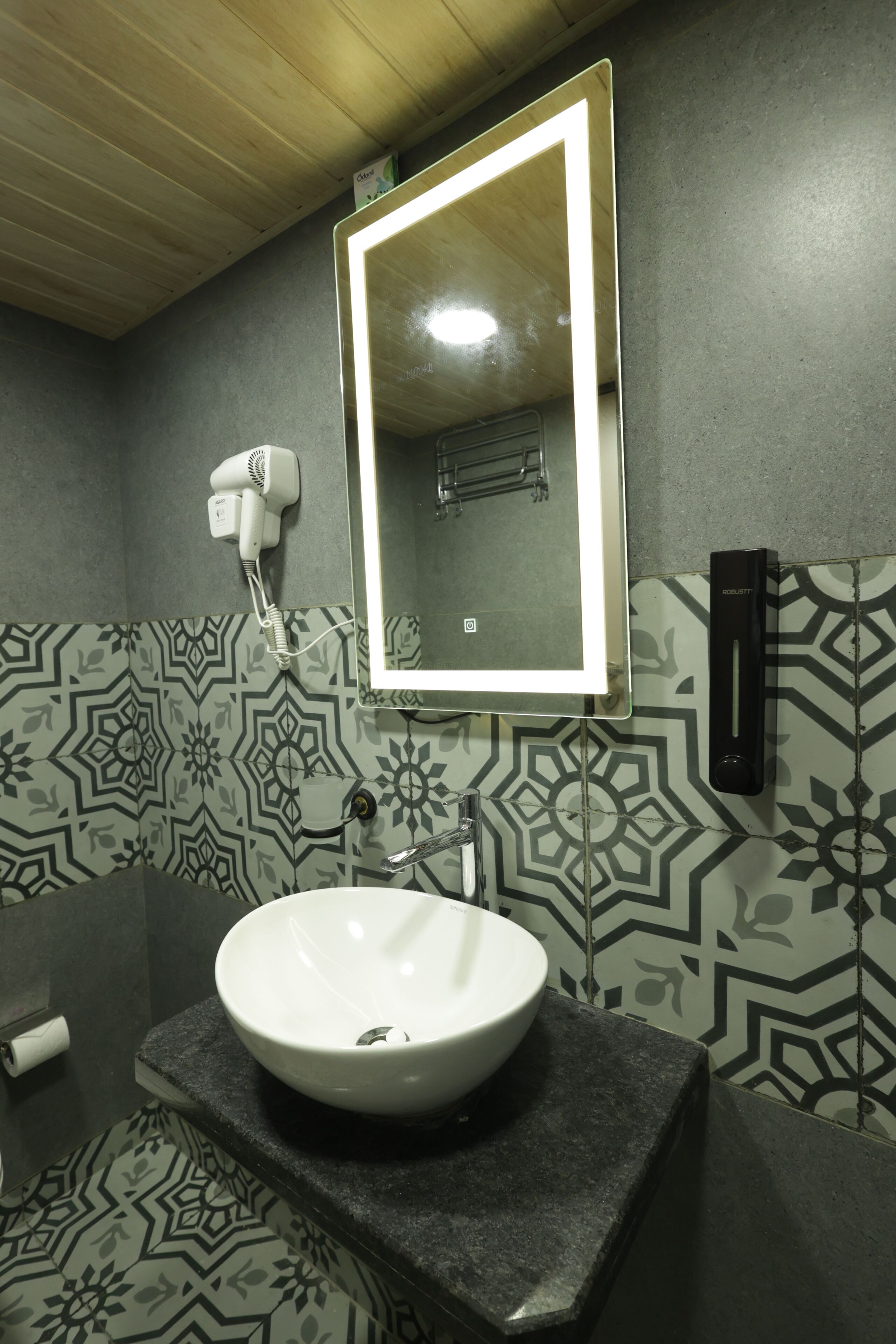 A bathroom with a white sink, a mirror with a light, patterned tiles, and a hairdryer on the wall.