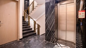 Un ascensor moderno con pantalla digital, pared de mármol con diseño geométrico y una escalera con puerta de madera y barandilla.