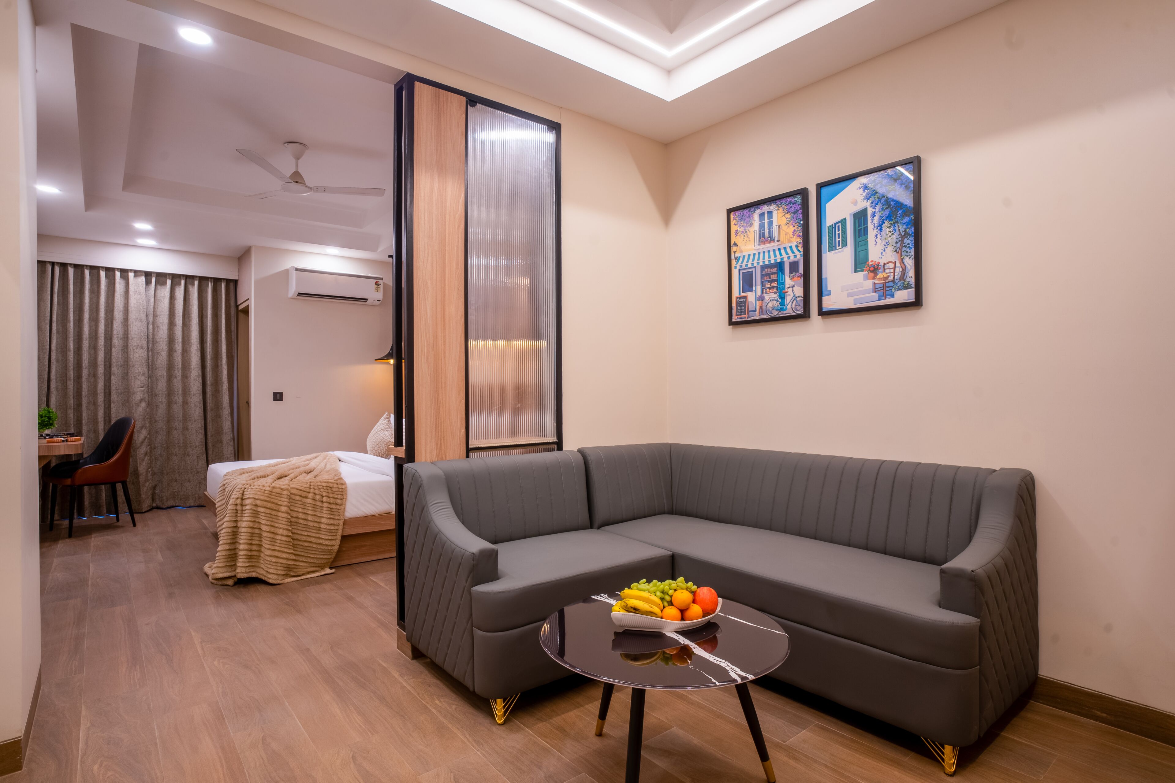Una sala moderna con un sofá seccional, una mesita redonda con fruta, una cama, un escritorio y un ventanal grande.