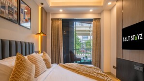 Habitación de hotel con cama, almohadas, televisión, ventana con cortinas y vistas al balcón.