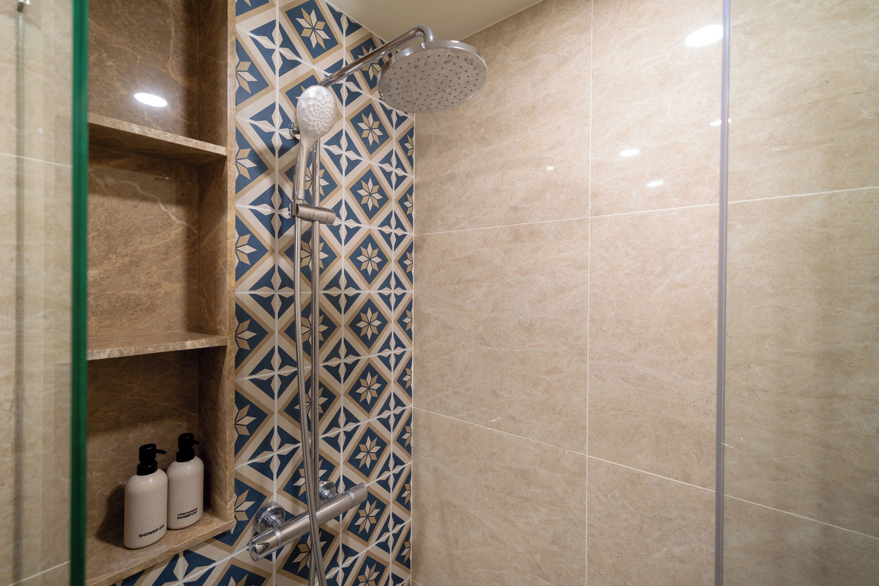 Een douche met een geometrisch tegelpatroon, een handdouche en twee flessen op een plank.