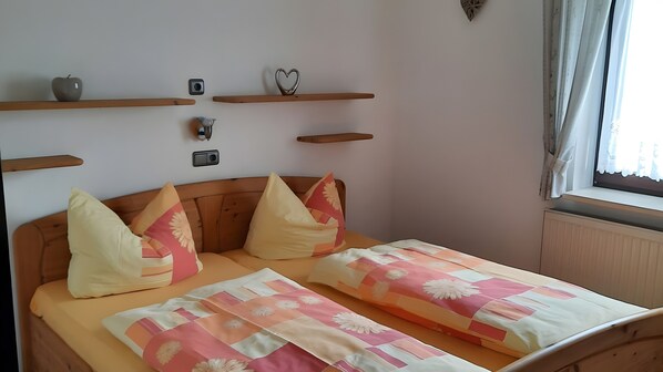 A bedroom with a wooden headboard, two beds with floral bedding, wooden shelves with decorative items, and a window with curtains.