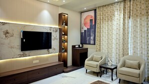 A modern living room with a flat-screen TV mounted on a marble wall, wooden shelving, two armchairs, a coffee table, and a large abstract painting.