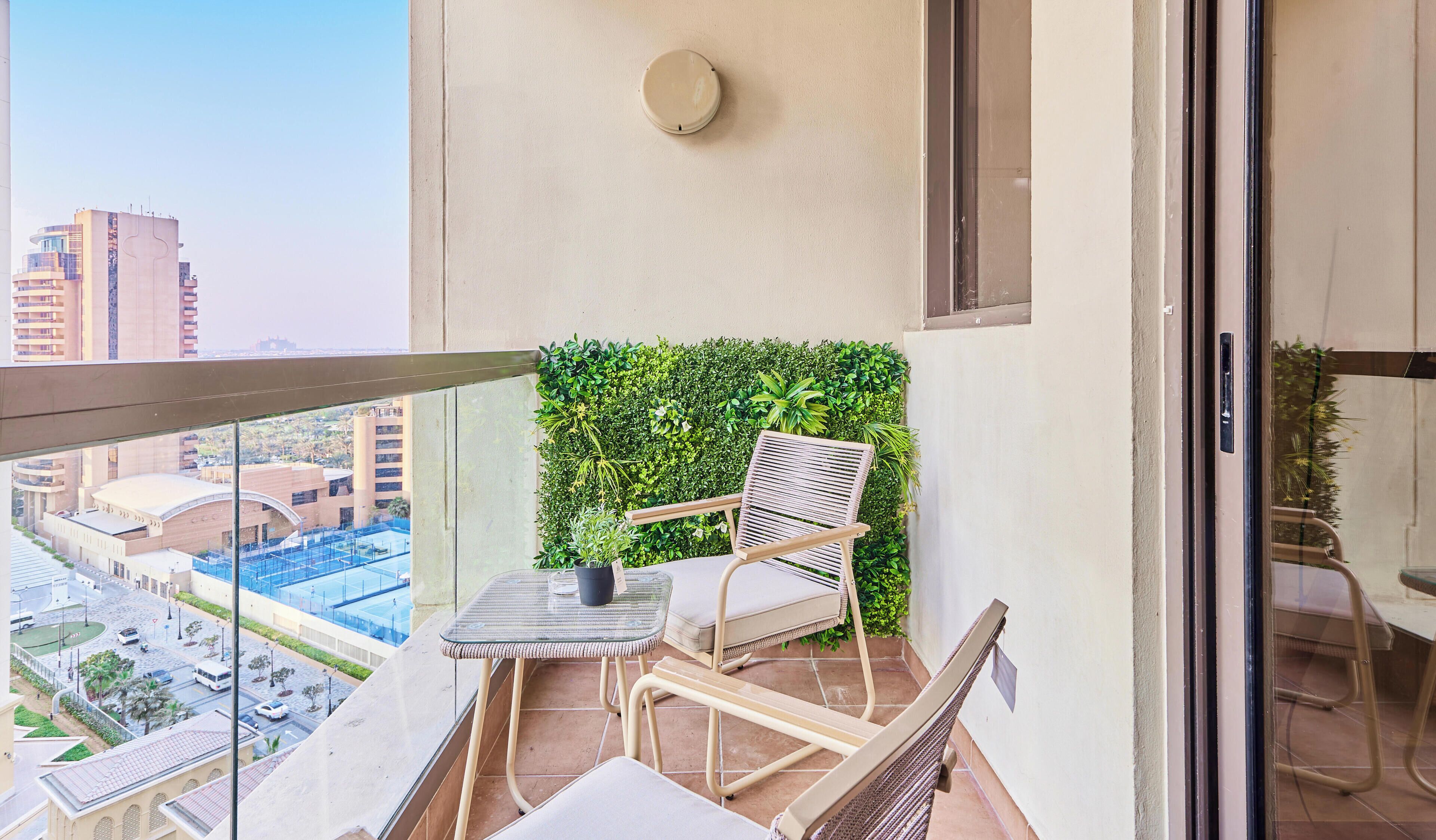 A balcony with a glass door, a small table with a chair, a green wall with plants, and a view of buildings.