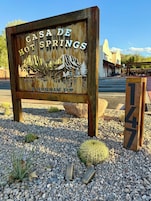 Un letrero rústico para Casa de Hot Springs con una ilustración de una montaña y un árbol, rodeado de grava y cactus.