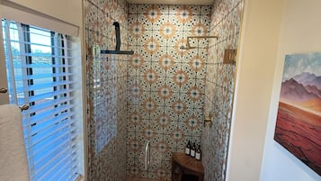 Eine Dusche mit gemusterten Fliesen, einer Glastür, einem Handtuch auf dem Boden und einem kleinen Tisch mit Flaschen.