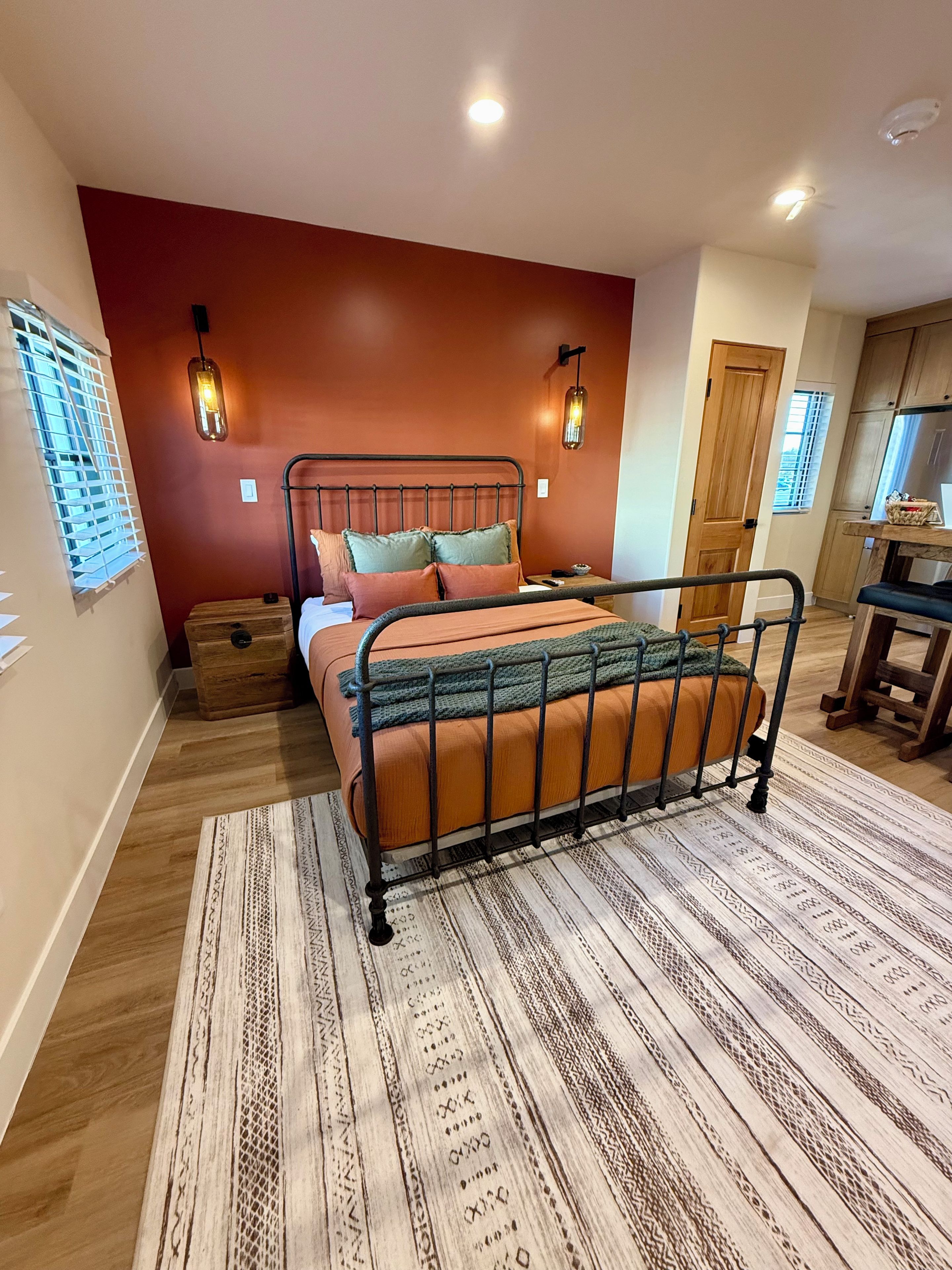 Un dormitorio con cama, mesitas de noche, ventana con persianas, piso de madera y una zona de cocina al fondo.