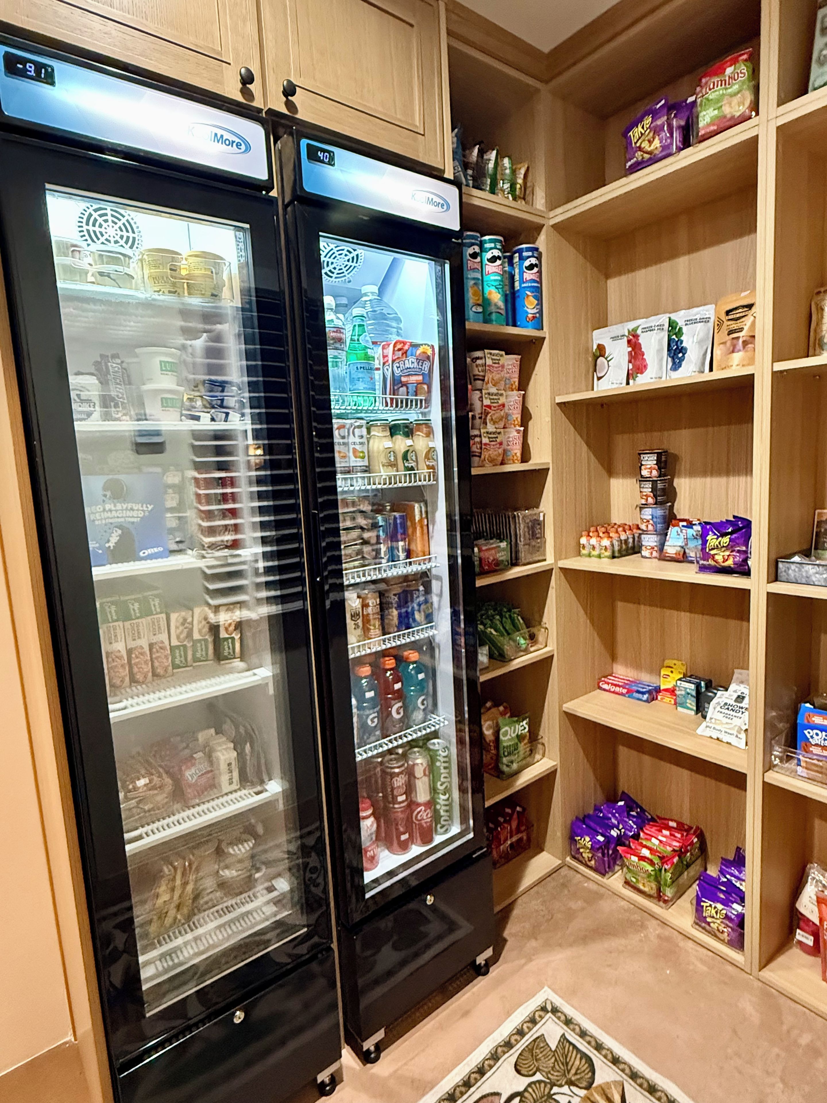 Een keuken met twee zwarte koelkasten vol drankjes en snacks, houten kasten en planken met diverse etenswaren.