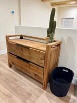 Un aparador de madera con un cactus encima, un cesto de basura de fieltro negro y una pared blanca con un termostato.