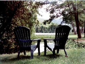 Deux chaises Adirondack noires, avec une petite table placée entre elles, sont disposées sur une pelouse, avec un lac et des arbres en arrière-plan.