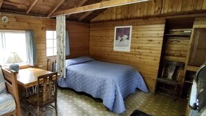 A wooden cabin room with a bed, a table, a chair, and a fireplace.