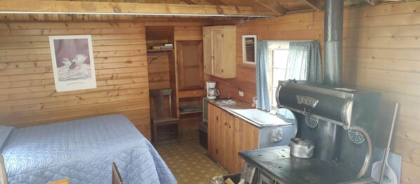 A cabin with a bed, a wooden cabinet, a stove, and a window with curtains.