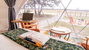 Ein Zelt mit einem Laptop auf einem Tisch, einem Bett mit floraler Tagesdecke und Blick auf Bäume sowie Stühle draußen.