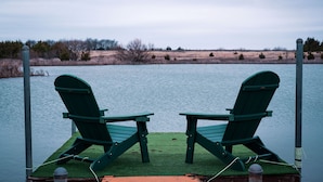 Twee groene Adirondack-stoelen op een steiger boven het water, met een grasrijke oever op de achtergrond.