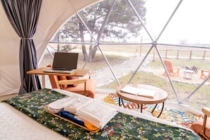 A tent with a laptop on a table, a bed with a floral bedspread, and a view of trees and chairs outside.