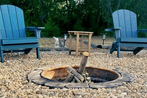 A fire pit with logs, surrounded by pebbles, with two Adirondack chairs and a wooden bench in the background.
