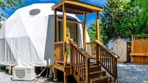 A dome-shaped tent with a wooden staircase leading to a covered entrance, surrounded by gravel and trees. - Unique Woodland Glamping Dome with Stargazing & Nature Retreat near Wolfe City, TX (Wolfe City)