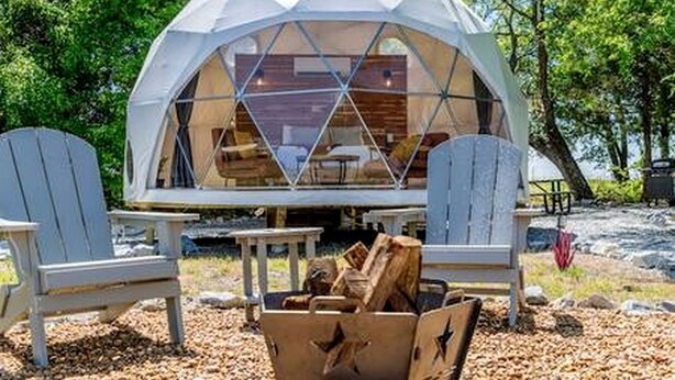 A geodesic dome structure with a wooden interior, two Adirondack chairs, and a fire pit. - Unique Woodland Glamping Dome with Stargazing & Nature Retreat near Wolfe City, TX (Wolfe City)