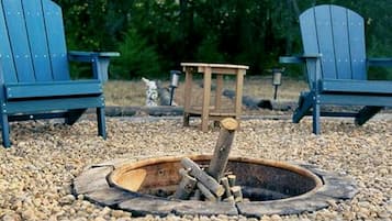 Un foyer avec des bûches, entouré de galets, avec deux chaises Adirondack et un banc en bois en arrière-plan.