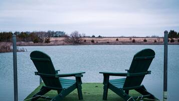 Two green Adirondack chairs on a dock over water with a grassy shoreline in the background.