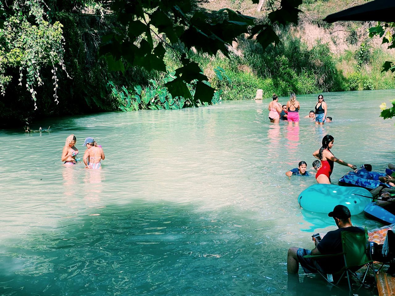 Un groupe de personnes se trouve dans un environnement naturel, près d’une étendue d’eau, avec des arbres et de la végétation en arrière-plan.