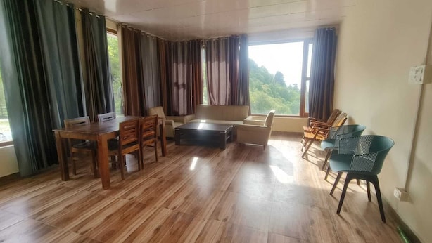 A room with a wooden dining table, chairs, a sofa, and a coffee table. There are curtains on the windows, and a view of greenery outside.