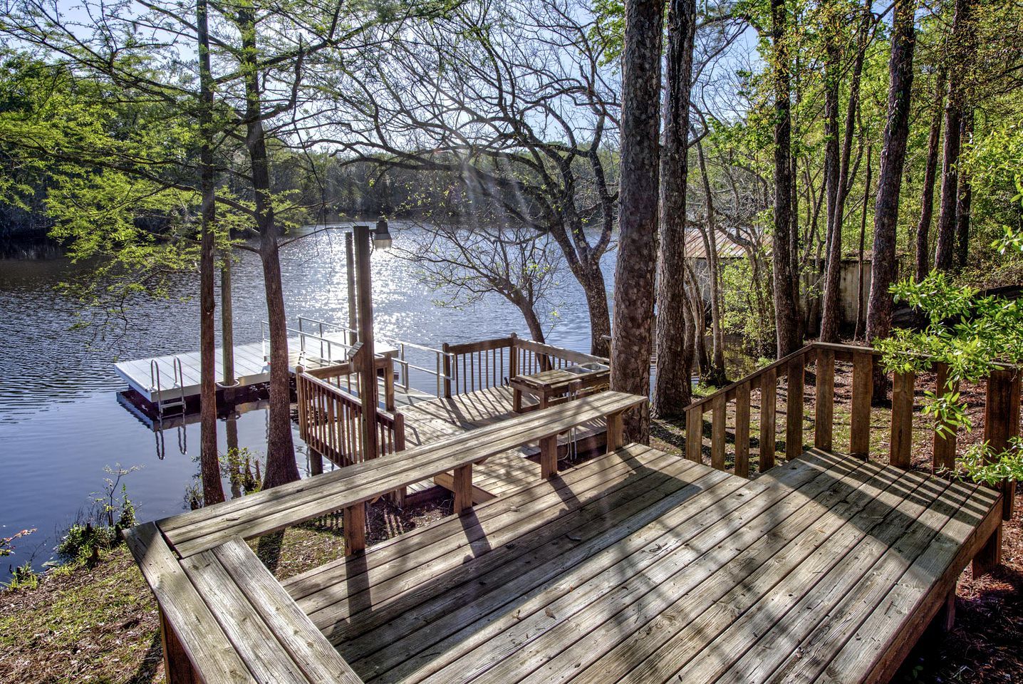 Une terrasse en bois avec un banc, un petit quai pour bateaux et une zone boisée avec des arbres et une étendue d’eau.