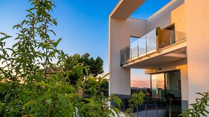Un edificio moderno de dos pisos con balcón, ventanas amplias y follaje circundante.