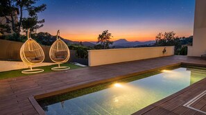 Una piscina en la azotea con dos sillones colgantes, una terraza de madera y vistas al atardecer sobre las montañas.