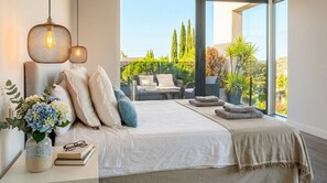 Un dormitorio con un ventanal grande, una cama con almohadas, una mesita de noche con un florero y libros, y vistas al exterior.