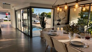 A dining area with a table set for two, a pool visible through glass doors, and a cactus plant.