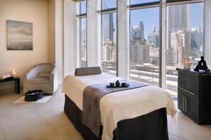 A bedroom with a bed, a chair, a small table with candles, and a view of the city through large windows. - Kempinski Central Avenue Residence  (Dubai)