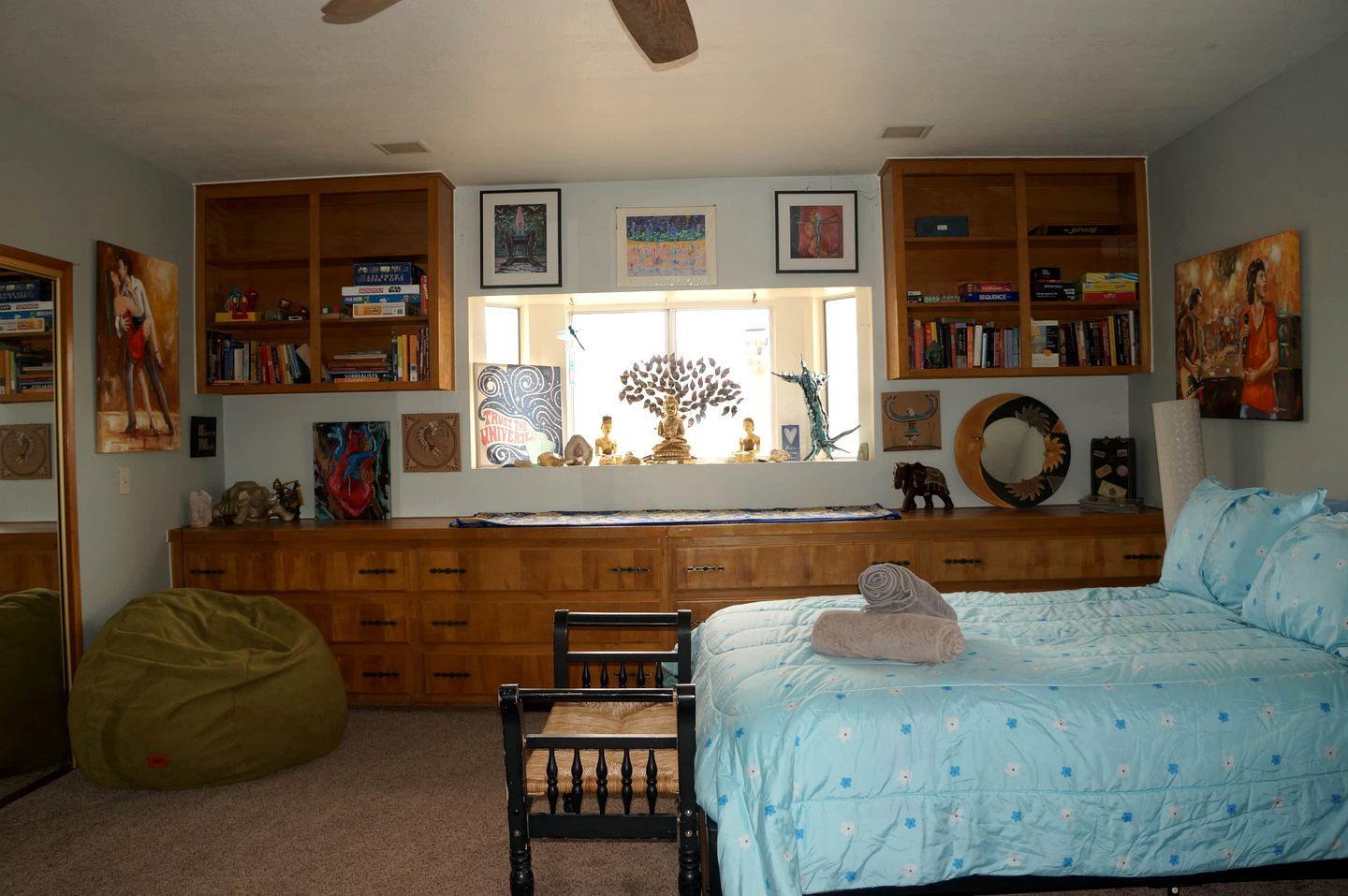 A bedroom with a bed, a desk, a chair, a bean bag, and bookshelves filled with books and decorative items.