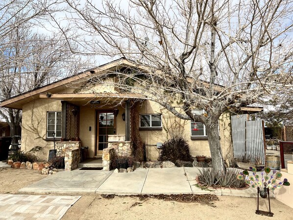 A single-story house with a sloped roof, a front porch, and a small front yard with plants and a tree.