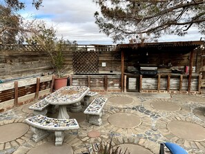 An outdoor patio with mosaic-tiled tables and benches, a barbecue grill, and a wooden fence.