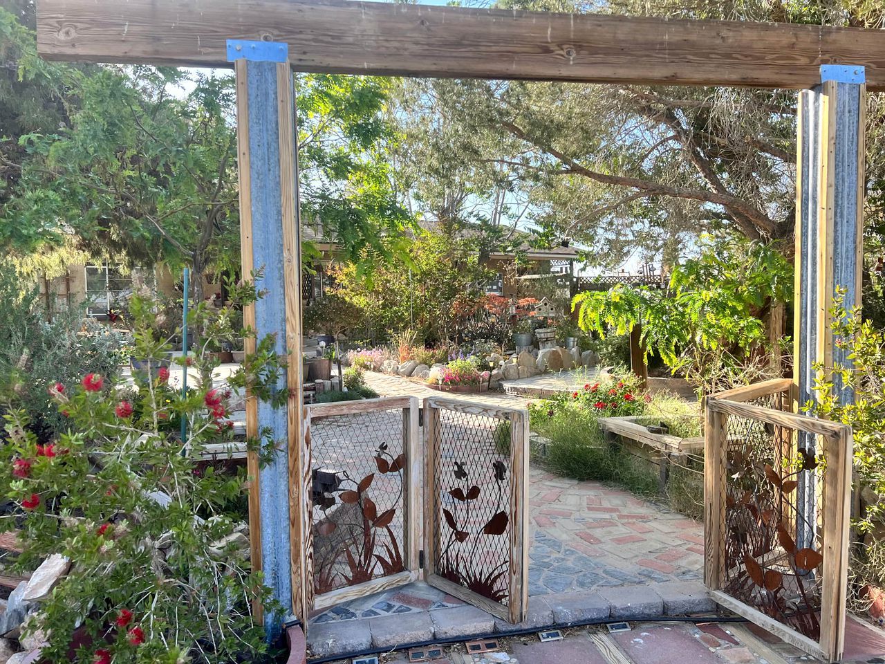 Un jardín con un porche de madera, una verja de metal y diversas plantas y flores.